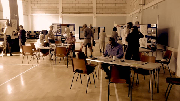 image of a hall containing several tables and chairs plus information boards showing laminated images. people are viewing the information boards, indicative of a community consultation