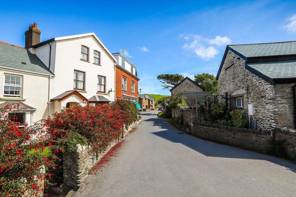 Photograph of a village street, looking slightly uphill. It's a sunny day, with bright blue sky. there is a stone building on the right and a white house on the left, with fuschia bushes in front. 